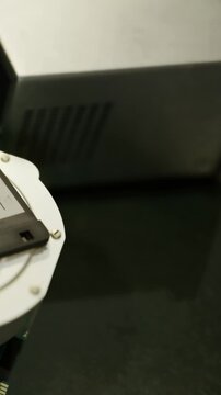 A computer monitor stands next to a small electronic device on a desk in an office setting. The wall is visible in the background, and the surface is dark.