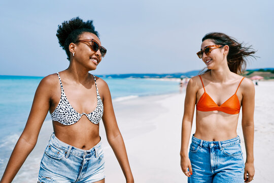 Two smiling young women, friends, walk along a sunny beach. They wear swimwear tops and denim shorts, enjoying their summer vacation by the clear blue ocean.