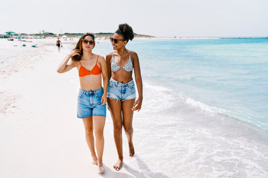 Two diverse young women, wearing swimwear and denim shorts, walk barefoot along a pristine white sand beach. They enjoy the clear turquoise ocean and sunny summer day together.