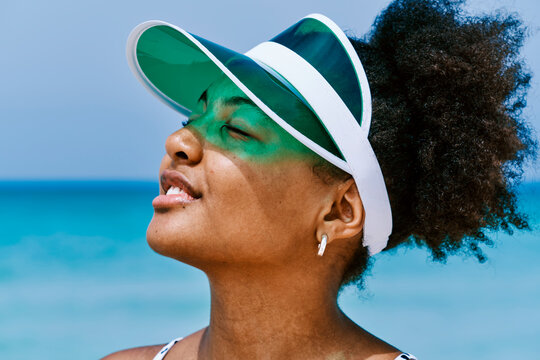 Young Black woman with an afro wearing a green visor, eyes closed, enjoying the sun and warmth on a bright day at the beach. The ocean is in the background.