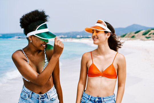 Two diverse young women walk and smile on a sunny beach. They wear swimwear tops, denim shorts, and colorful visors, enjoying their summer vacation.