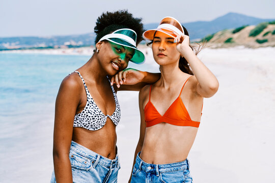 Two young women, friends, stand on a sunny beach. One smiles, resting her head on the other's shoulder. They wear vibrant visors, swimwear tops, and denim shorts, enjoying their summer vacation.