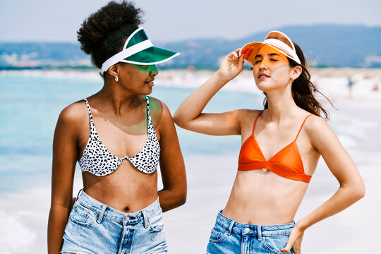 Two diverse young women on a sunny beach. One in a green visor looks at her friend. The other adjusts her orange visor, eyes closed, enjoying the sun. They wear swim tops and denim shorts.