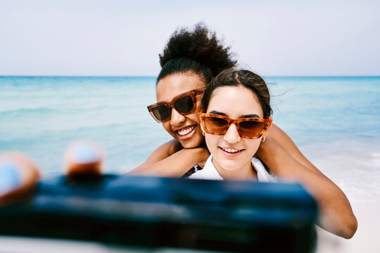 Two happy young women in stylish sunglasses are taking a selfie together on a beautiful beach. The clear blue ocean provides a stunning backdrop for their summer vacation.