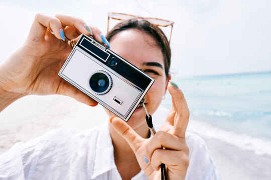 A young woman with colorful nails holds a retro film camera, aiming it at the viewer. She smiles while enjoying a sunny day at the beach, capturing vacation memories.