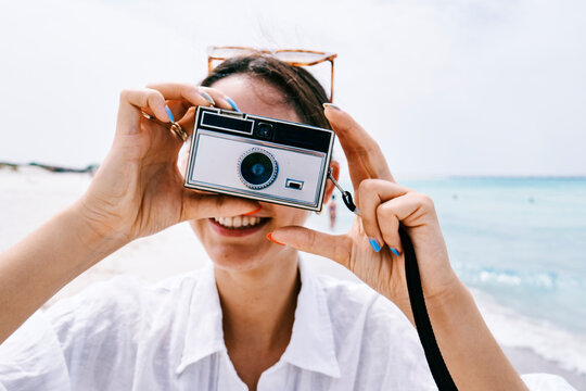 A young woman smiles while holding a vintage camera to her face, capturing a moment on a beautiful sunny beach. The blue ocean stretches behind her.