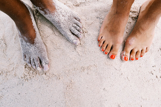 Two pairs of feet on a sandy beach. One person has dark skin and sand-covered feet. The other has lighter skin with bright orange painted toenails. Relaxing by the ocean.