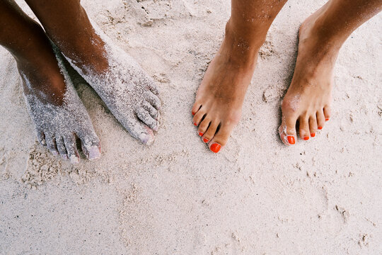 High angle view of two individuals standing on white beach sand. One person has feet covered in fine sand while the other displays vibrant orange nail polish on their toes.