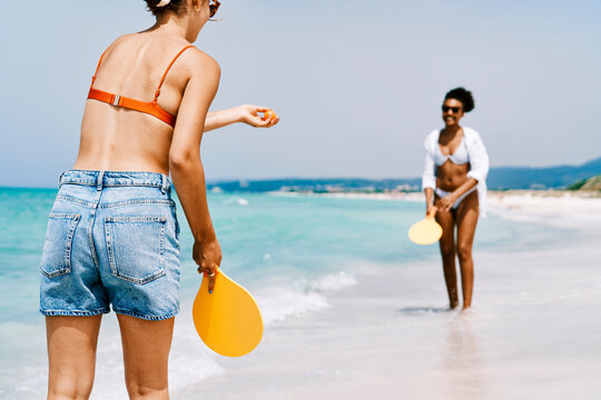 Two women play paddleball on a sunny beach. One woman holds a paddle and ball, ready to serve. The other stands in the distance, smiling. The turquoise ocean provides a beautiful backdrop.