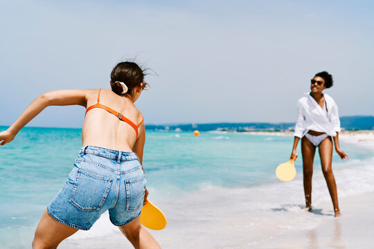 Two young women play beach tennis on a sunny sandy beach with turquoise water. One woman hits the orange ball with a yellow paddle, while the other prepares to return it. Summer fun.