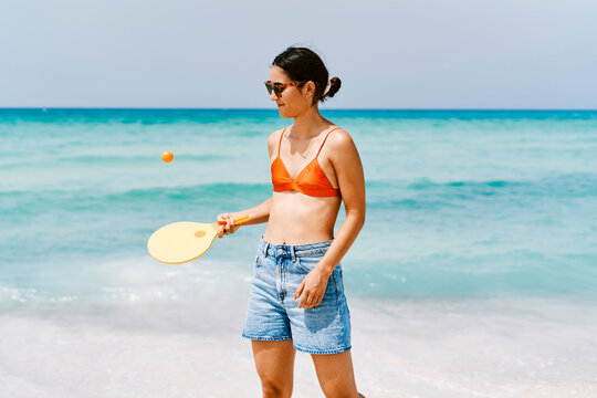 A young woman in sunglasses, an orange bikini top, and denim shorts stands on a sunny beach. She holds a yellow paddle, ready to hit an orange ball. The turquoise ocean is behind her.