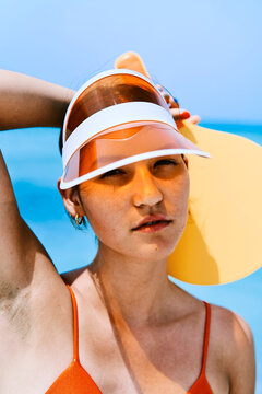 Young woman with freckles wears an orange visor and swimsuit. She holds a yellow paddle against a bright blue sky, looking directly at the viewer.