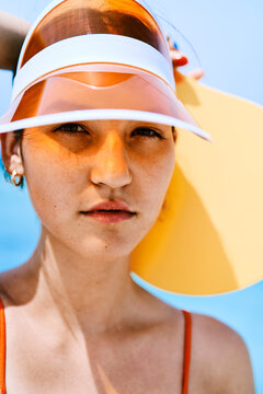 Close-up portrait of a young woman wearing an orange transparent visor and a red swimsuit. She looks directly at the camera with a calm expression under bright sunlight.