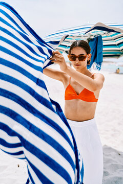 A young woman in sunglasses and an orange bikini top holds a blue and white striped towel flowing in the breeze on a sunny beach with umbrellas.