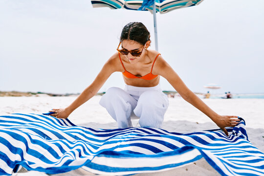A young woman wearing an orange bikini top and white trousers squats on the white sandy beach to spread out a blue and white striped towel under a striped umbrella.