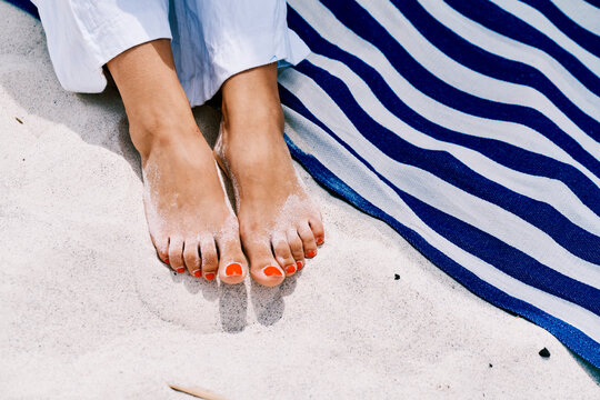 A person's feet with orange nail polish are covered in sand, resting on a beach next to a blue and white striped towel. This evokes relaxation and summer vacation.