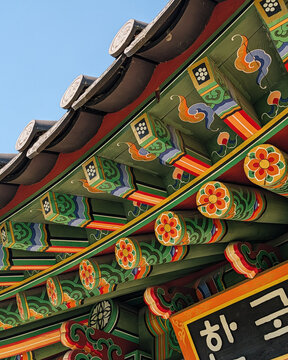 Traditional korean dancheong coloring on roof eaves. Close-up of wooden roof eaves with decorative coloring of vibrant green, red, and orange floral patterns under a clear blue sky. 