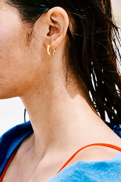 A woman's ear with a gold hoop earring. Her wet hair and skin indicate she just came out of the water. She is wearing a red swimsuit strap and a blue towel.
