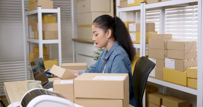Asian woman small business owner scanning parcel box barcode beside laptop while e-commerce packaging workflow continues showing online shopping parcel preparation for customer delivery process