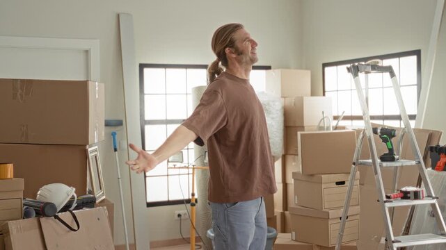 Man standing with arms outstretched beside ladder and cordless drill amid packed boxes in building; satisfaction renovation.