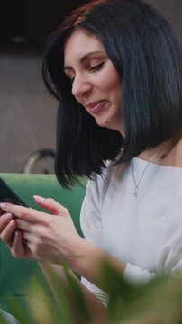 Portrait of attractive woman sitting on sofa and texting message on smartphone on background modern kitchen at home. Thoughtful young woman using cellphone at domestic kitchen. Slow motion