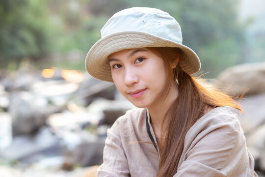 Beautiful young Asian woman wearing light bucket hat smiling peacefully sitting near rocky river stream enjoying tranquil nature scenery relaxing outdoors during pleasant daytime holiday trip