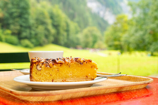 A slice of traditional Swiss nut cake on a white plate. The cake is served on a tray outdoors with a blurred forest and mountain background in Switzerland.