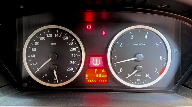 A car's dashboard instrument cluster shows silver-ringed dials for the speedometer and tachometer, plus illuminated red warning lights and a central orange digital information display.