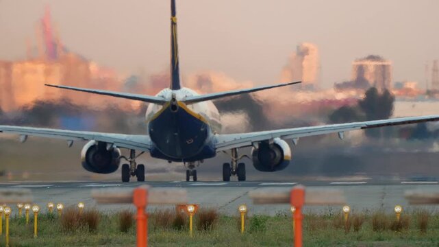 Commercial passenger jet accelerates down airport runway takes off. View from behind plane with engine heat haze against a hazy city skyline during a warm sunset sunrise. Travel transport concept