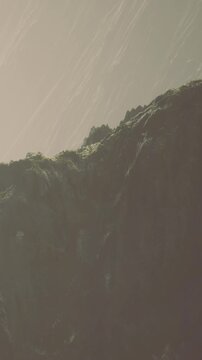 Clouds cover the sky as rain falls in the distance. A large rock formation rises above the ground. The scene shows natural elements interacting with weather conditions.