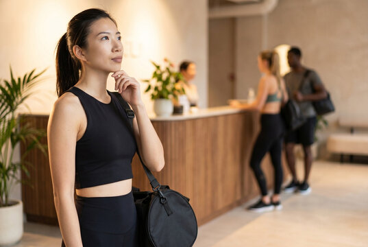 Asian woman in sportswear holding gym bag, contemplating her workout at the fitness studio