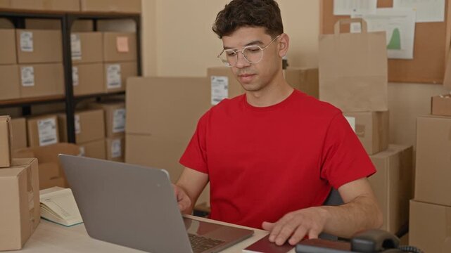 Man smiling, holding passport and pumping fist beside laptop and stacked parcels at packing desk in building; entrepreneurial pride.