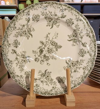 Vista de cerca de un plato con decoraci&oacute;n floral cl&aacute;sica, con patr&oacute;n de flores y hojas y ramillete floral en el centro, en expositor de madera en estante de comercio
