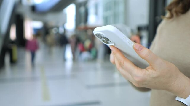 Hand holds smartphone in public space. Person walks through bright indoor corridor. Background shows blurred people and light-colored walls. Focus remains on device throughout clip