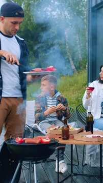 Pretty satisfied loving black-haired woman drinking red wine and enjoying time with her sons which eating pizza when her husband prepare red pepper and tasty sausages on barbecue for family party in