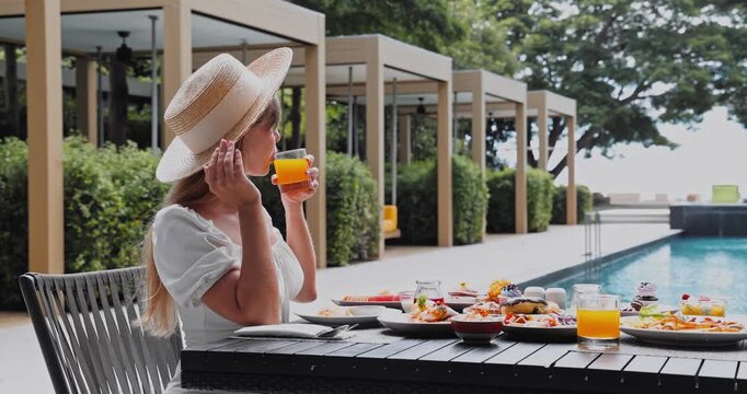 Woman drinking orange juice at dining table, outdoor luxury resort patio beside swimming pool with cabanas, summer vacation wellness, hotel breakfast service, upscale travel marketing