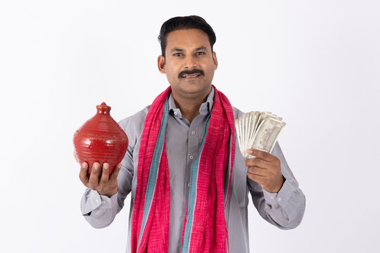 Portrait of smiling indian farmer in white topi holding indian rupees and piggy bank while standing against white background