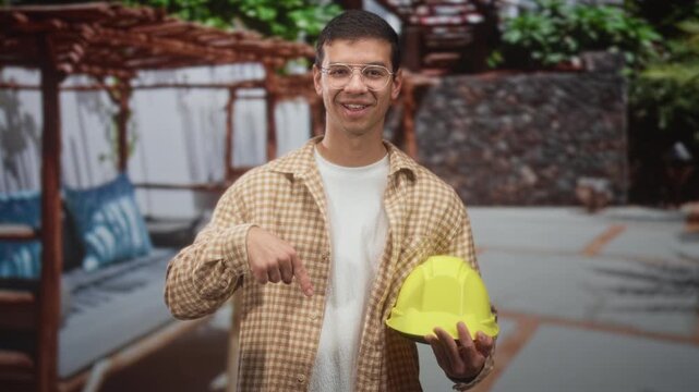Man points finger at a yellow hardhat while holding the helmet in one hand inside a building interior, smiling and wearing safety glasses; safety confidence.