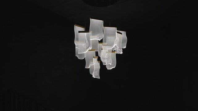 Modern lighting fixture made of white fabric like elements hangs and gently rotates against a dark wall