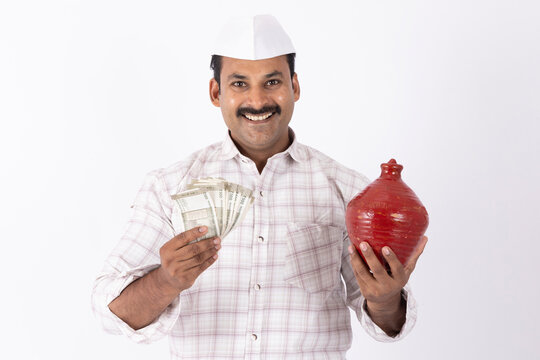 Portrait of indian farmer man in maharashtrian topi holding indian rupees and piggy bank while standing against white background