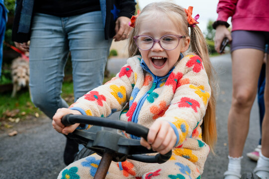 Girl with ponytails riding a toy car with her family walking behind her