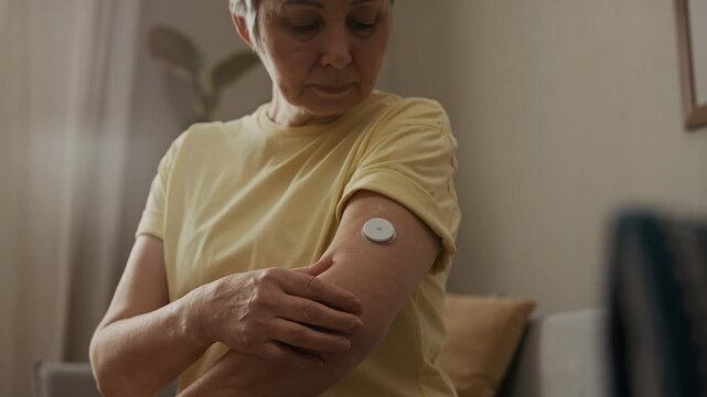 Cropped close up shot of senior woman checking glucose sensor applied to upper arm while monitoring sugar level with device at home, slow motion