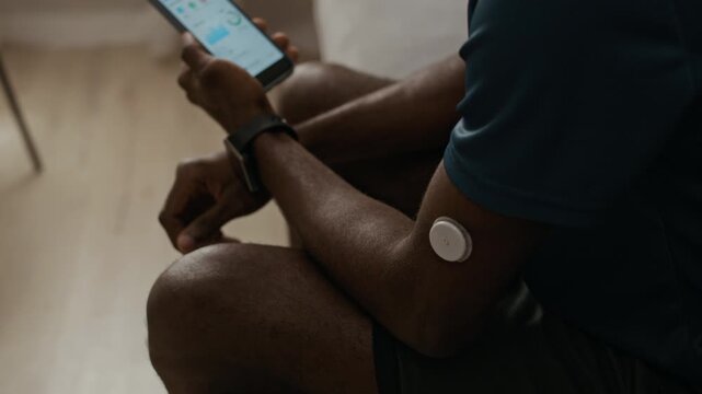 Over shoulder shot of young adult Black man viewing CGM data on mobile app connected to glucose sensor on upper arm while measuring blood sugar level at home, tilt down camera movement
