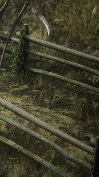 Wooden fence made of logs stands in a grassy field. A curved scythe leans against the fence. The time is late afternoon and the grass is dry and brown.