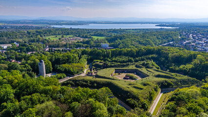 Obraz premium Panoramic aerial view of Fort Prussia in Nysa, showcasing geometric grass-covered bastion fortifications, a stone tower, the vast Nysa Lake, and a mountain range on the horizon in bright daylight.