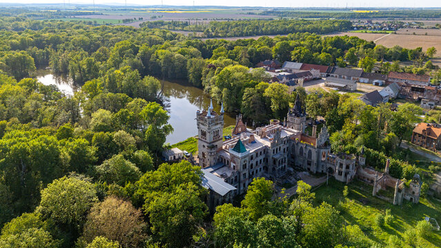Wide aerial panorama of the neo-gothic Kopice palace ruins, featuring the outbuildings complex, a pond, and a vast park, with distant farmlands under a blue sky in sunny afternoon light.