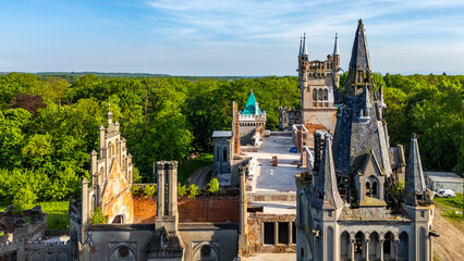 Wide drone shot of the Kopice palace ruins, neo-gothic architecture with numerous towers, carved details, and brick walls, surrounded by a vast green forest and park on a sunny day. © Adam