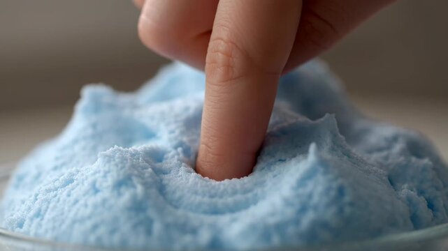 Finger pressing into soft blue foam creating an indentation. Playful texture exploration with vibrant color.
