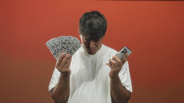 Man holding fanned dollars and smartphone, hands visible, furrowing brow while comparing cash to phone in studio; financial confusion.