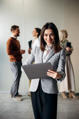 Fototapeta premium Confident businesswoman smiling, holding a laptop, standing in an office with colleagues in the blurred background